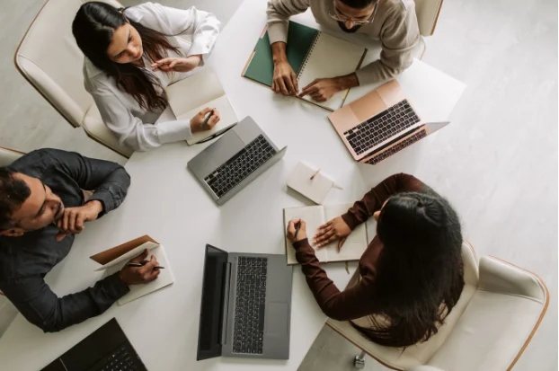 Vista cenital de cuatro profesionales trabajando alrededor de una mesa blanca con portátiles y cuadernos